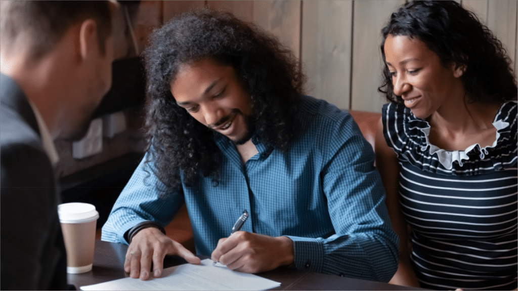 A couple is seated at a table, signing a rental agreement with a landlord. The man, with curly hair and a beard, is smiling as he signs the document, while the woman, with curly hair and wearing a striped top, looks on with a smile. The landlord, partially visible, leans in slightly, holding the document steady. A coffee cup is placed on the table next to them. The setting appears warm and informal, suggesting a positive and agreeable interaction. This image aligns with the blog post content on finding and keeping great tenants through clear communication and setting expectations.