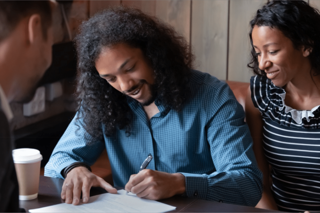 A couple is seated at a table, signing a rental agreement with a landlord. The man, with curly hair and a beard, is smiling as he signs the document, while the woman, with curly hair and wearing a striped top, looks on with a smile. The landlord, partially visible, leans in slightly, holding the document steady. A coffee cup is placed on the table next to them. The setting appears warm and informal, suggesting a positive and agreeable interaction. This image aligns with the blog post content on finding and keeping great tenants through clear communication and setting expectations.